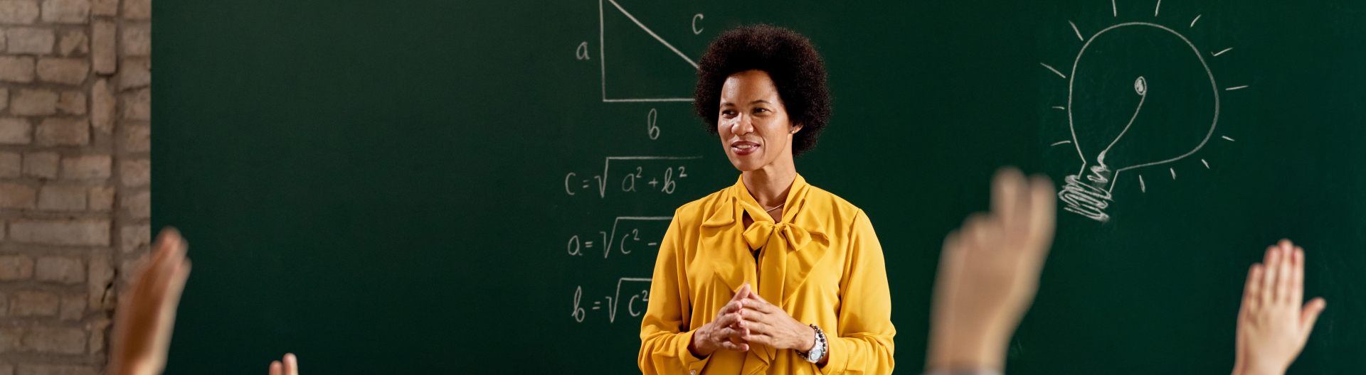 Active Teacher Header featuring a female, African American teacher standing in front of a chalk board with math formulas and a light bulb written on it. Students are raising their hands to answer a question.