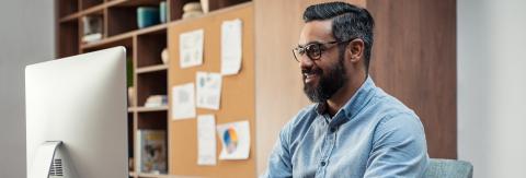 Photo of a man with a beard and glasses, wearing a blue shirt, sitting at a desk and smiling at his desktop computer screen.