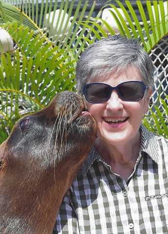 A woman posing outdoors, wearing sunglasses, and a sea lion is kissing her on the cheek. 