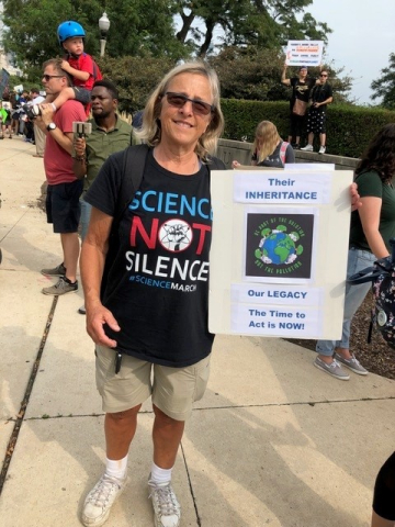 Retiree Dorelle Ackermann holding a sign that says "Their Inheritance. Our Legacy. The time to act is NOW!" She is outside at a protest, she is wearing a black shirt, shorts, and sunglasses.